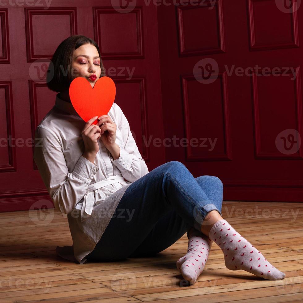 kortharige vrouw met gesloten ogen en lippen die kus vormen, zittend op de vloer, benen gekruist met hartvorm voor haar kin foto