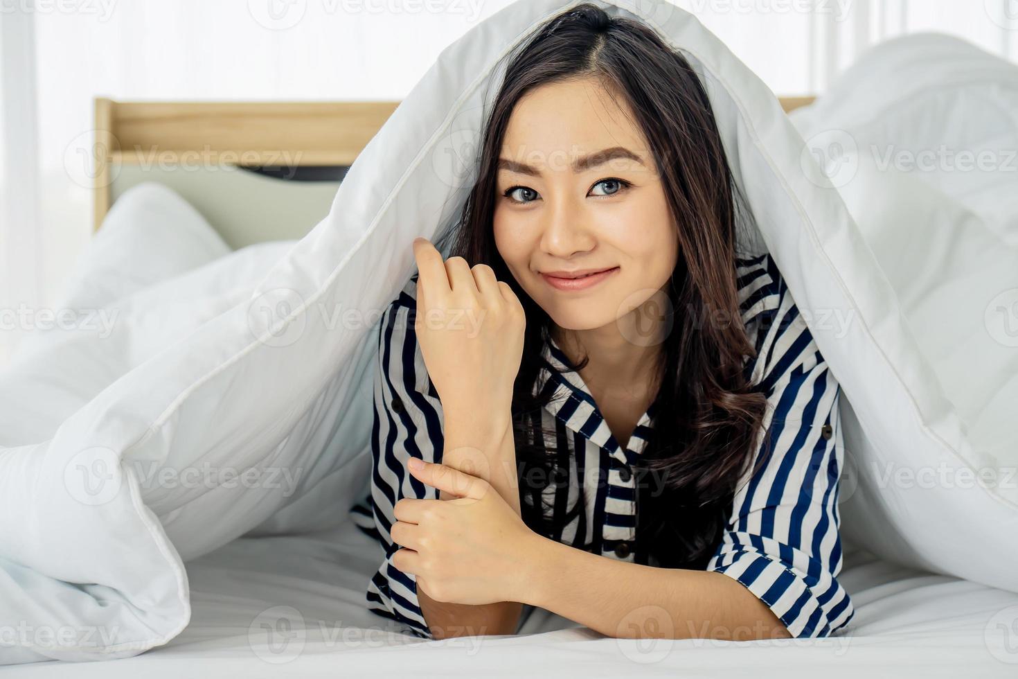 jonge aziatische vrouw die 's ochtends wakker wordt, glimlachende vrouw die onder een dekbed in haar slaapkamer rust. close-up portret van een mooie jonge vrouw en onder de deken. fijne goedemorgen foto