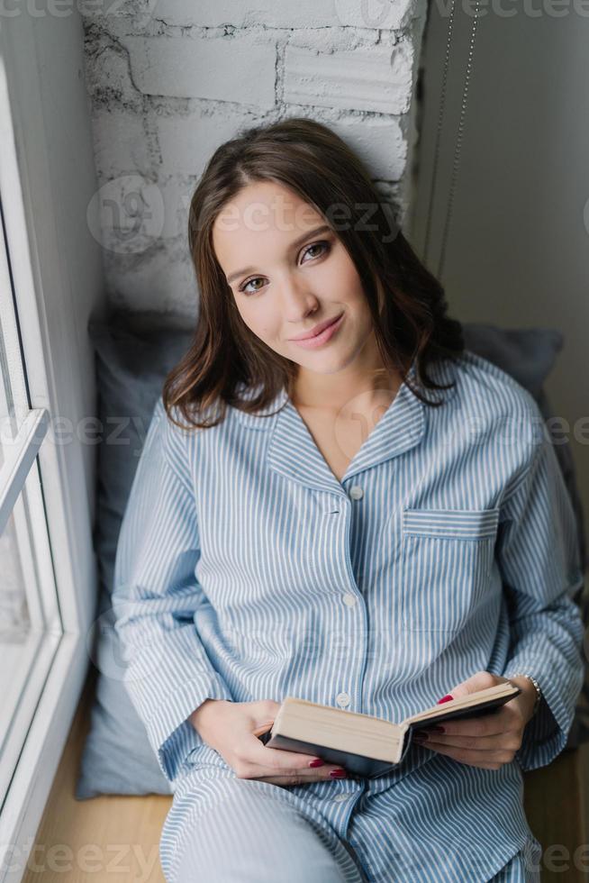 mooie ontspannen vrouw met zorgeloze uitdrukking, gekleed in nachtkleding, leest 's ochtends een boek op de vensterbank, geniet van vrije tijd, houdt van haar hobby. verticaal schot van tienermeisje met literatuur foto