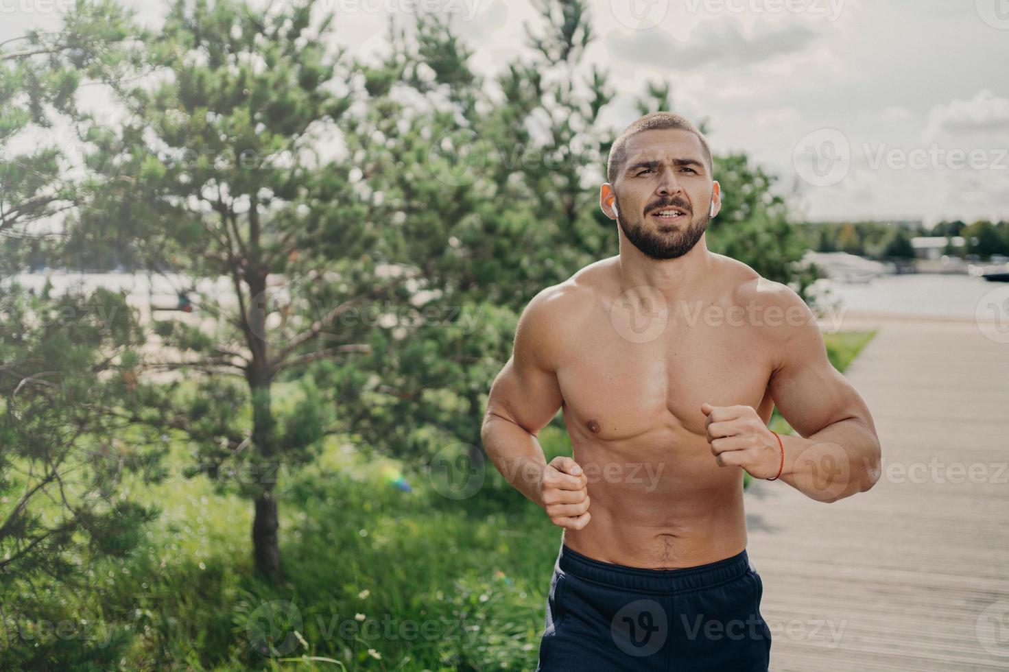 gezonde sterke bebaarde man hardloper heeft joggingoefening in de open lucht, ademt diep en luistert naar muziek in oortelefoons, heeft naakte torso, sterke gespierde armen. ochtendtraining en actief levensstijlconcept foto