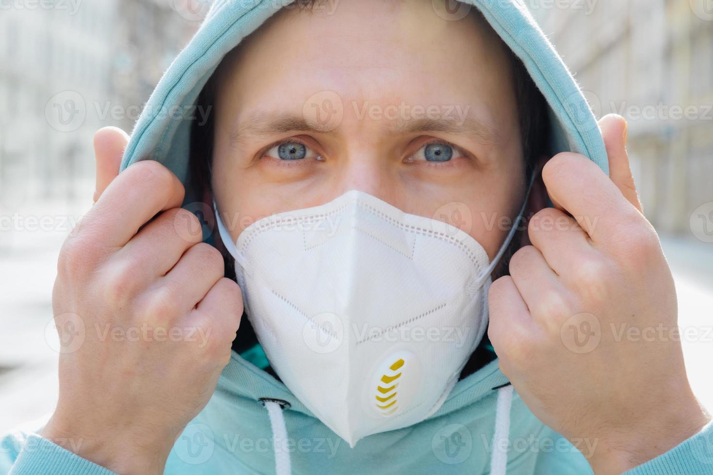 close-up shot van europese man beschermt gezicht met ademhalingsmasker, draagt hoodie, poseert buiten op straat, kijkt serieus naar camera, voorkomt coronavirus of covid-19, griep. straat tijdens quarantaine foto