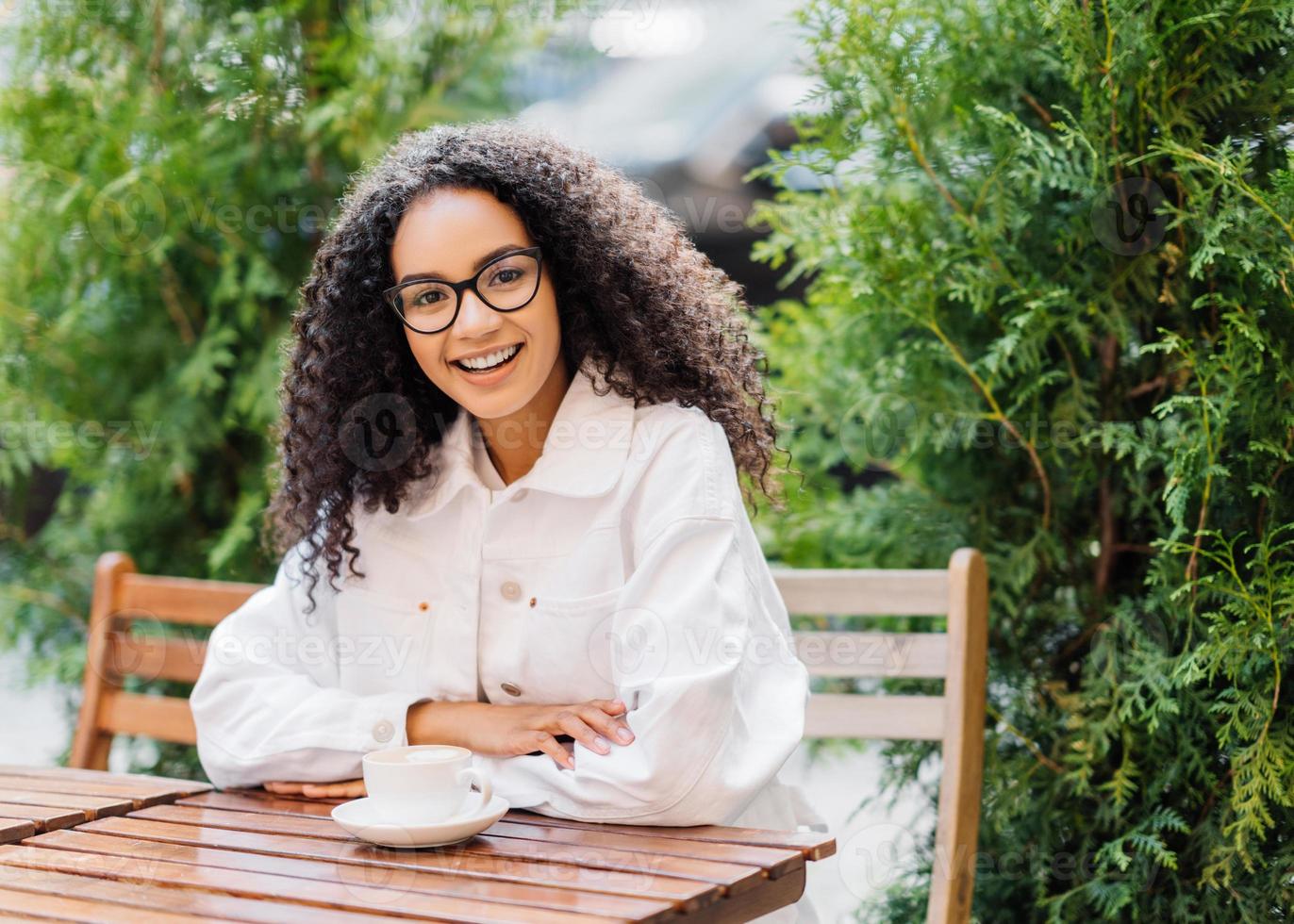 ontspannen vrolijke afro-vrouw in witte kleding, drinkt koffie in de buitencafetaria, zit aan houten tafel op stoel tegen groene thuja, heeft een gelukkige uitdrukking. mensen, levensstijl, vrije tijd concept foto