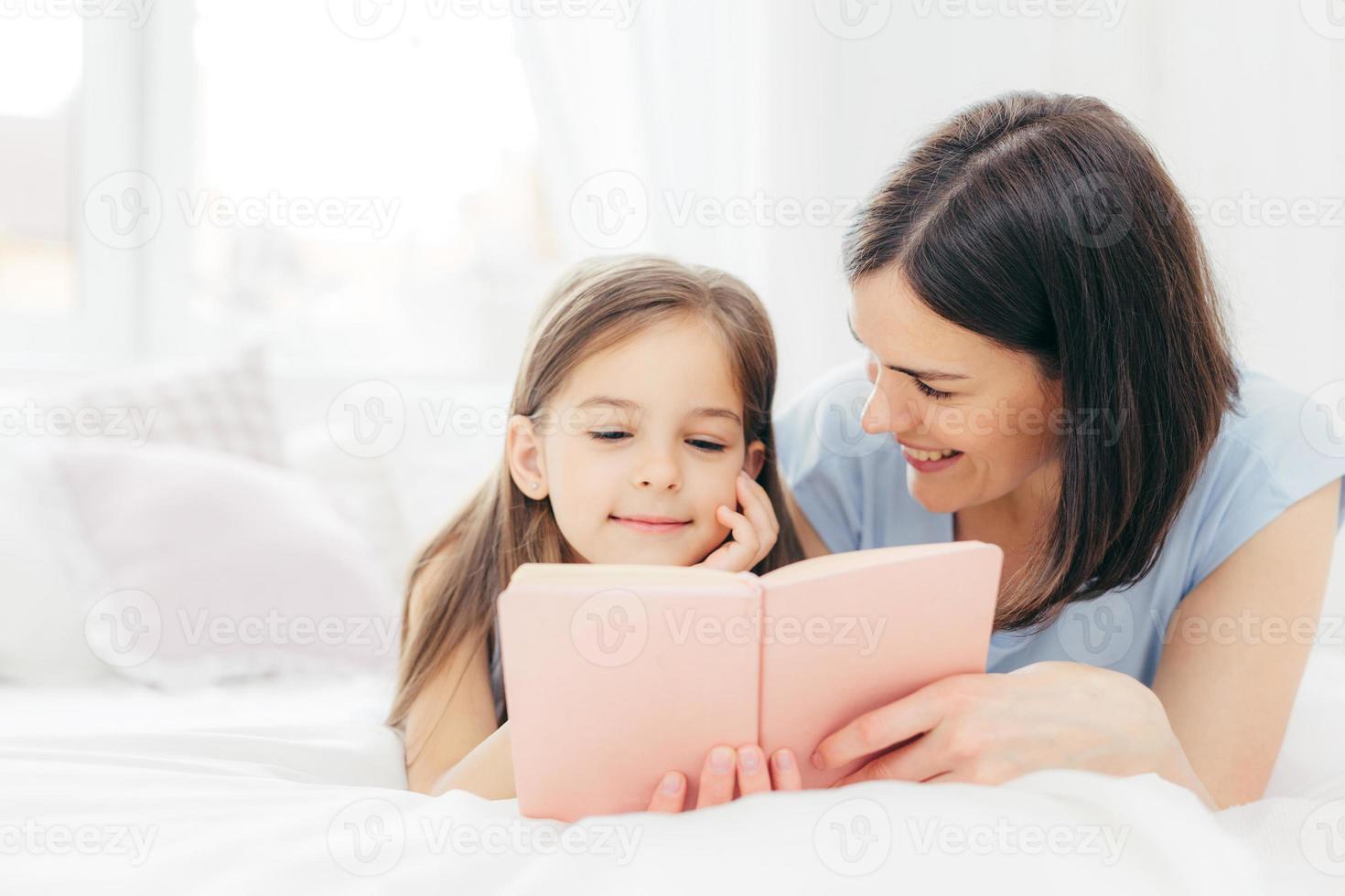 aanhankelijke jonge moeder met donker haar, kijkt blij naar haar kleine dochter, leest samen een interessant boek of sprookje, geniet thuis van de huiselijke sfeer. mensen, familie, jeugdconcept foto