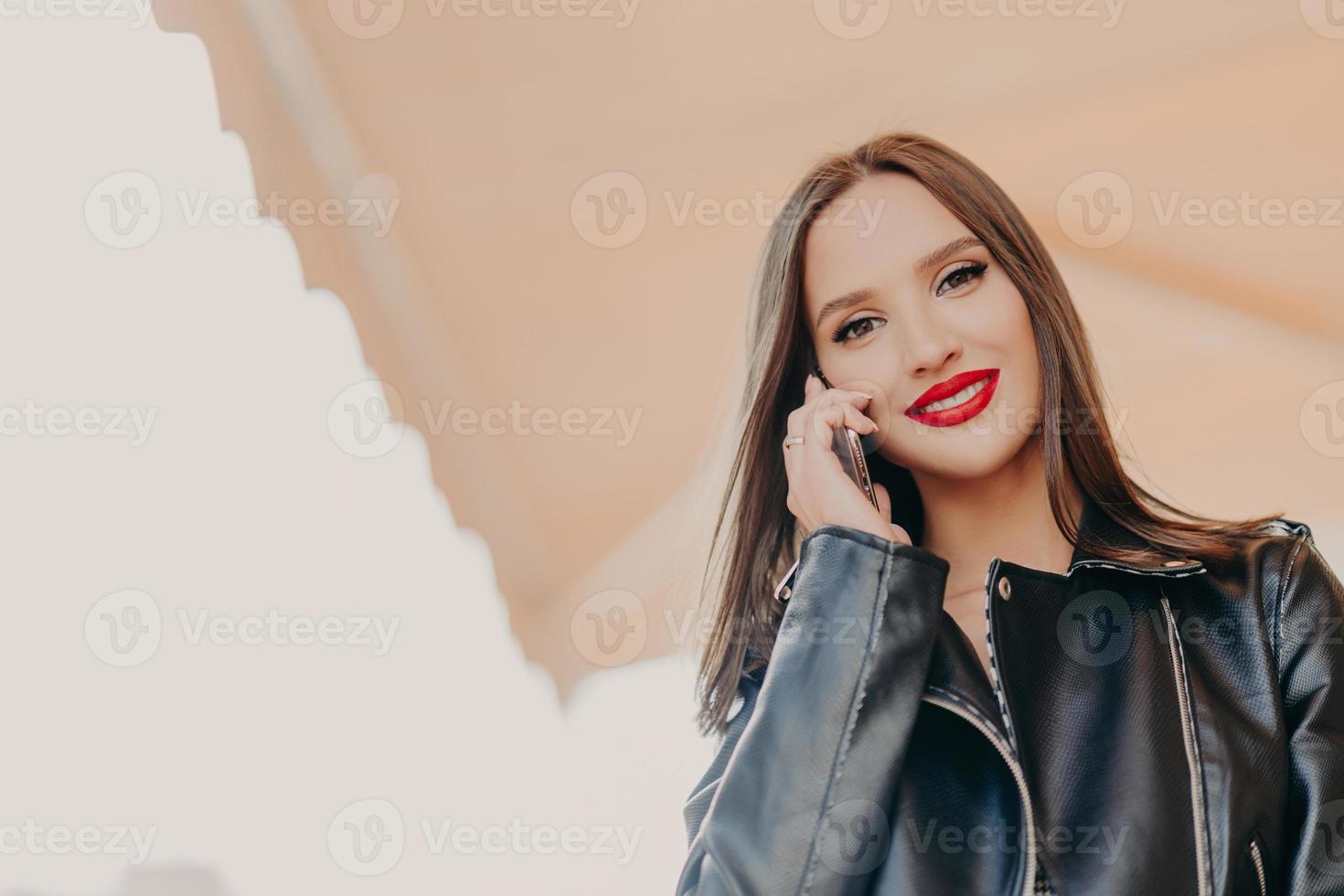 shot van mooie jonge duizendjarige vrouw praat graag met vriendje, draagt leren jas, heeft make-up, rode lippenstift, gebruikt moderne mobiele telefoon voor communicatie. mensen en gespreksconcept foto