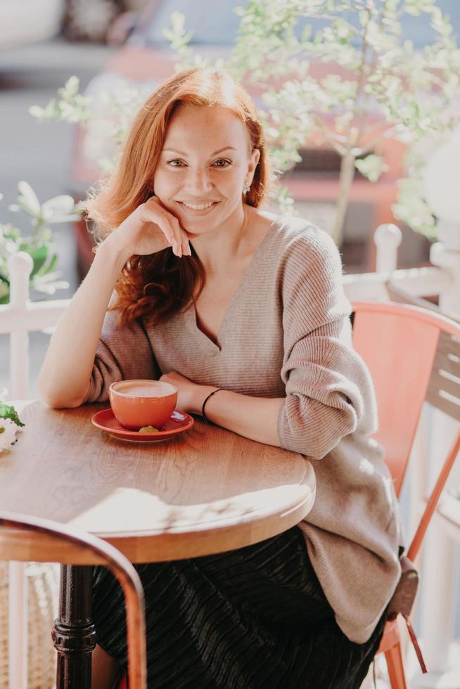 verticaal schot van vrolijke foxy vrouw gekleed in modieuze kleding, drinkt koffie, poseert in het terras, geniet van vrije tijd, heeft een aangename glimlach op het gezicht, houdt de hand onder de kin. levensstijl concept foto