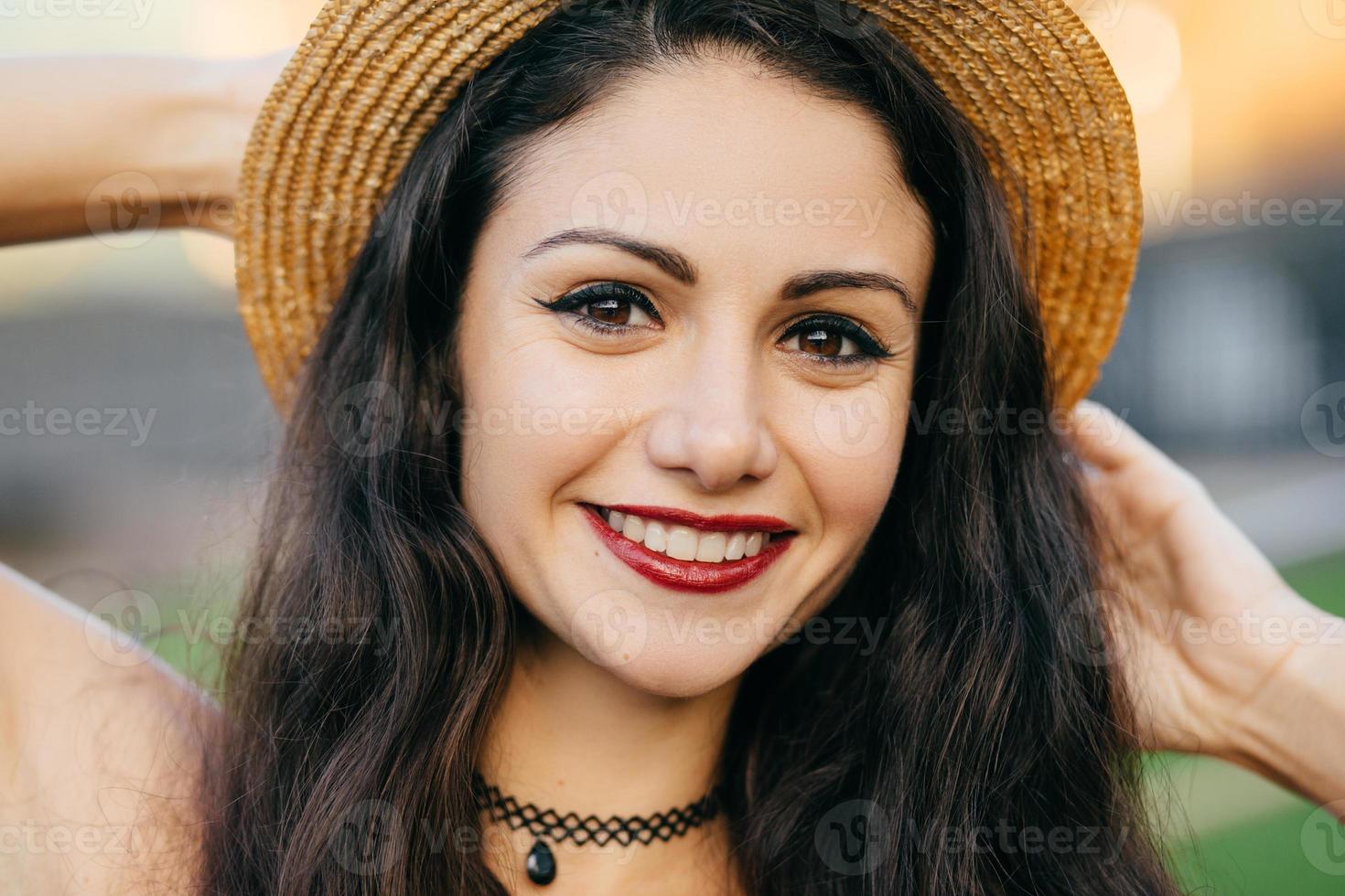 close-up portret van brunette vrouw met donkere ogen, zuivere huid en rode lippen met make-up met strohoed terwijl ze buitenshuis poseert. modieuze dame met een heerlijke uitdrukking en een aantrekkelijke glimlach foto