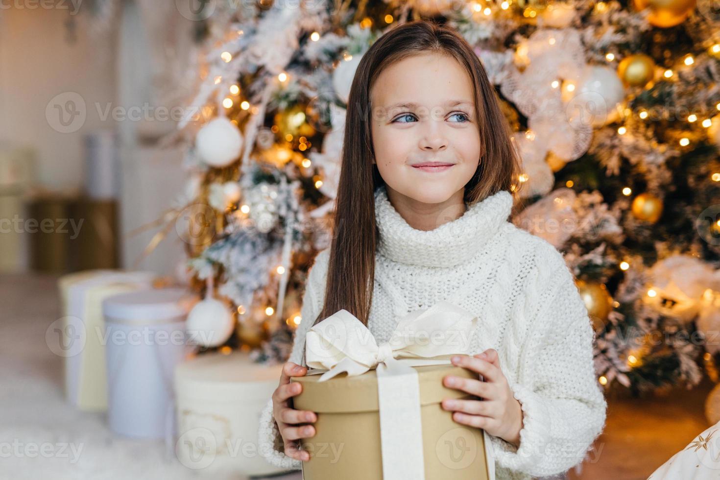 schattig lief klein kind met charmante uitstraling, blij om kerstcadeau te ontvangen, kijkt opzij met een gelukkige uitdrukking, heeft intrgue wat het heden is, poseert in de buurt van een prachtig versierde dennenboom. foto