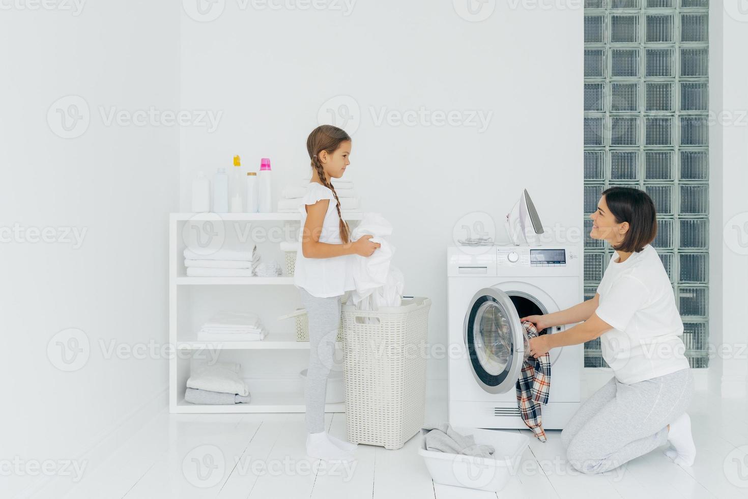 drukke brunette vrouw laadt de wasmachine met vuile kleren, haar dochtertje helpt, staat in de buurt van de mand en sorteert de was. gelukkige moeder en kind in de wasruimte. huishoudelijk werk concept. foto
