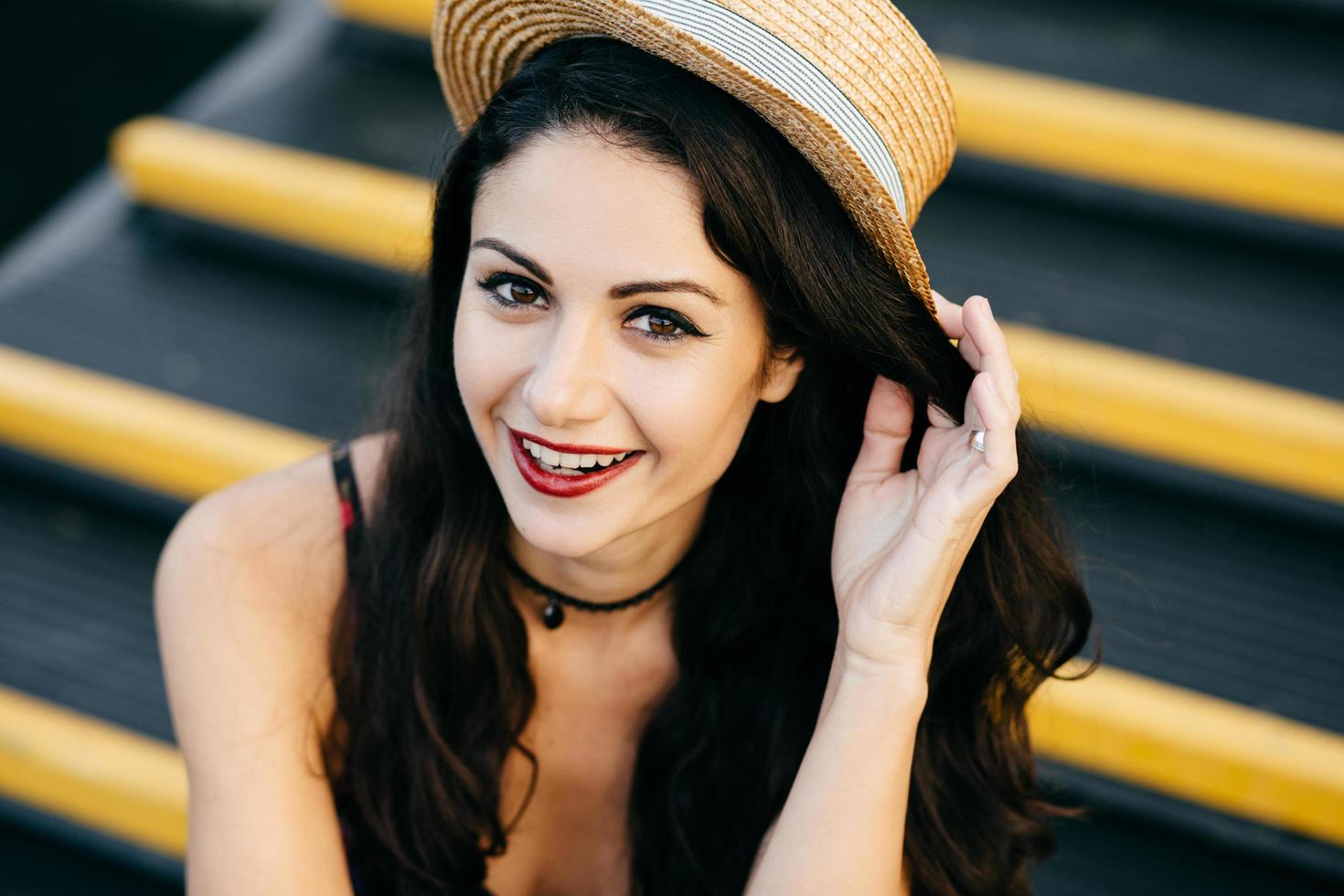 positieve vrouw met donker haar, glanzende ogen en rode lippen met een strohoed en ketting die zich ontspannen voelt en een goed humeur heeft terwijl ze op de trap zit. mensen, levensstijl, jeugd, modeconcept foto