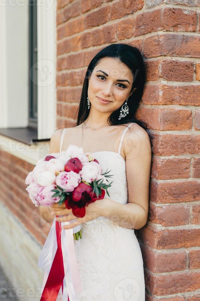 aantrekkelijke vrouwelijke bruid draagt witte trouwjurk, houdt boeket vast, staat in de buurt van bakstenen muur, heeft een aantrekkelijk uiterlijk. elegante jonge vrouw met make-up, heeft charmante glimlach viert haar huwelijk foto