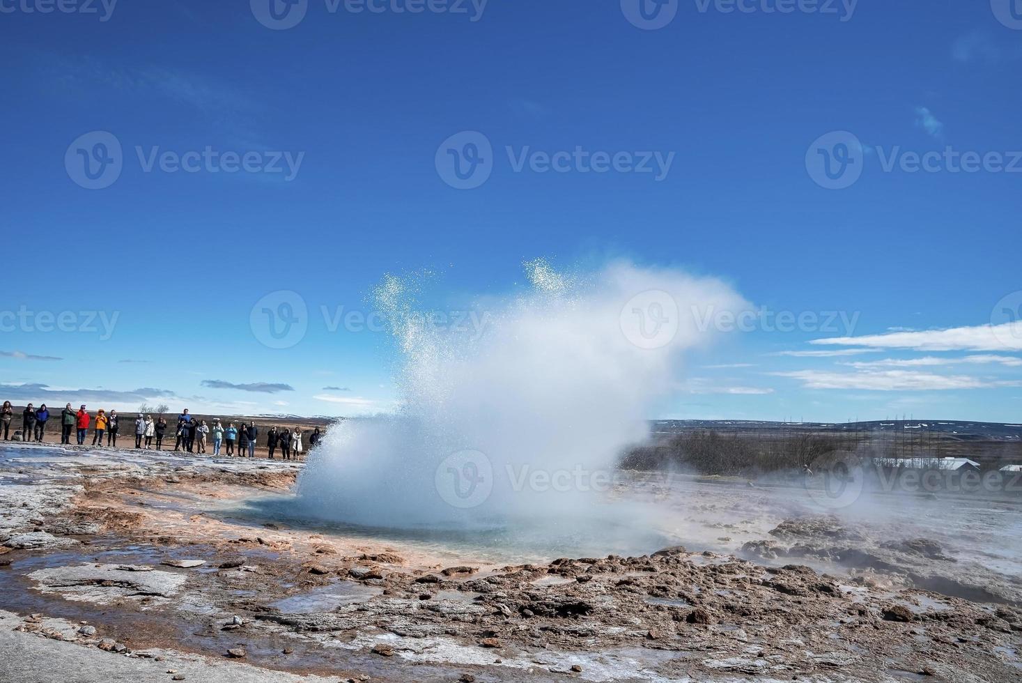 toeristen kijken naar de uitbarsting van de Strokkur-geiser in de vallei tegen de blauwe lucht foto
