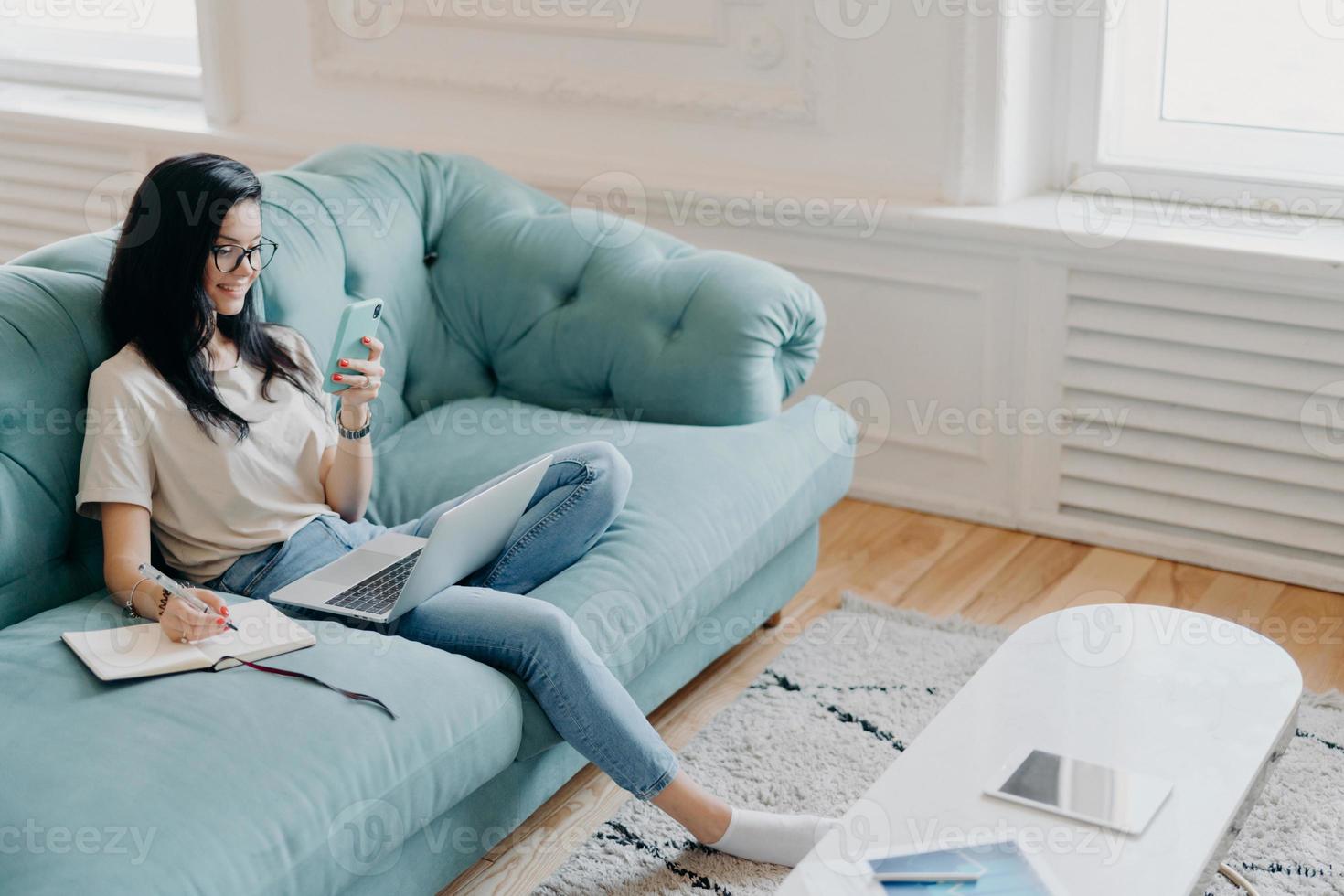 boven schot van mooie brunette vrouw gefocust op samrtphone-display, schrijft noodzakelijke informatie in notitieboekje, kijkt naar trainingswebinar, poseert in gezellige kamer op comfortabele bank, werkt vanuit huis foto