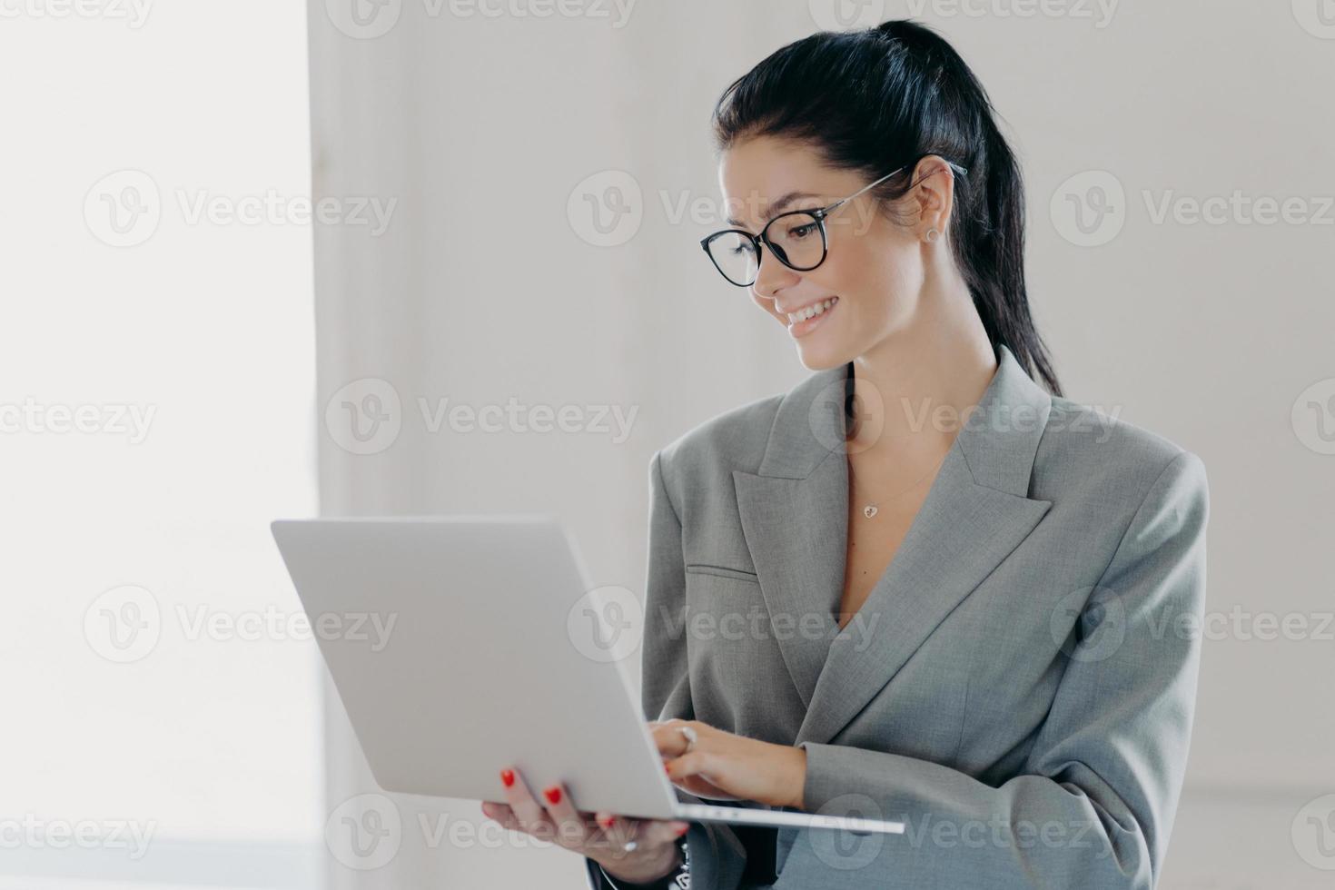 zelfverzekerde vrolijke vrouw econoom analyseert rapport over bedrijfsinkomsten op laptopcomputer, deelt multimediabestanden, heeft donker haar in paardenstaart gekamd, gekleed in formele outfit, staat binnen foto