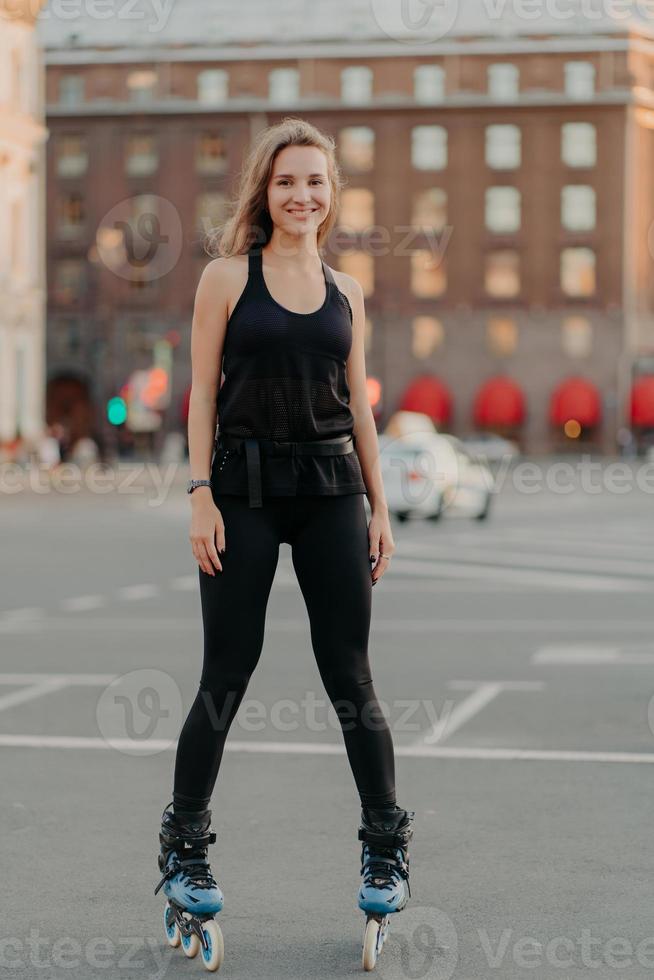 positief jong vrouwelijk model in sportkleding rijdt op bladen geniet van vrijetijdsactiviteiten poses op stedelijke plaats tegen onscherpe achtergrond staat in volle lengte. actieve levensstijl en skateconcept foto