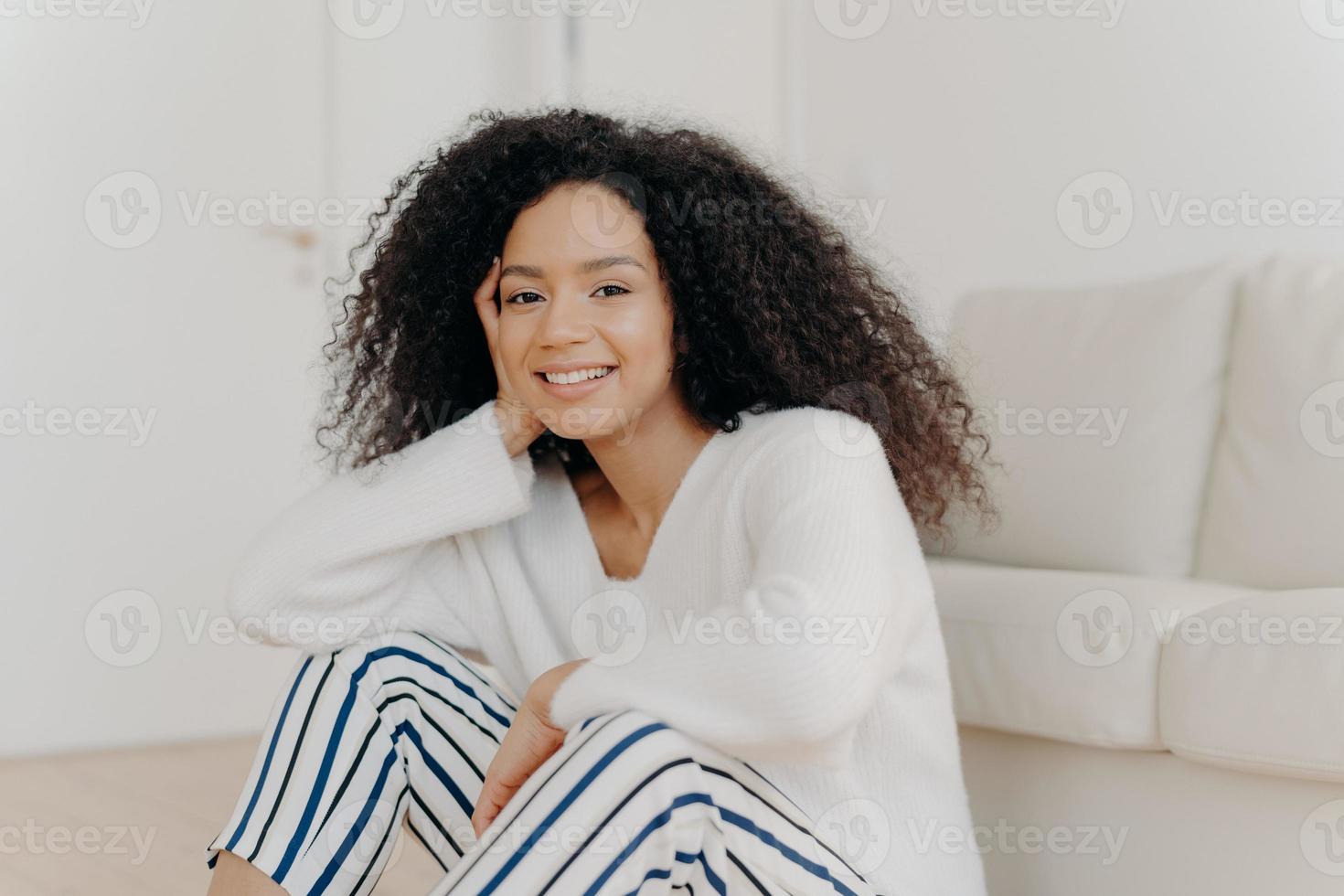foto van ontspannen opgetogen Afro-Amerikaanse vrouw met krullend kapsel, glimlacht zachtjes naar de camera, draagt een witte trui en gestreepte broek, zit op de vloer in de buurt van de bank in de woonkamer van een modern appartement