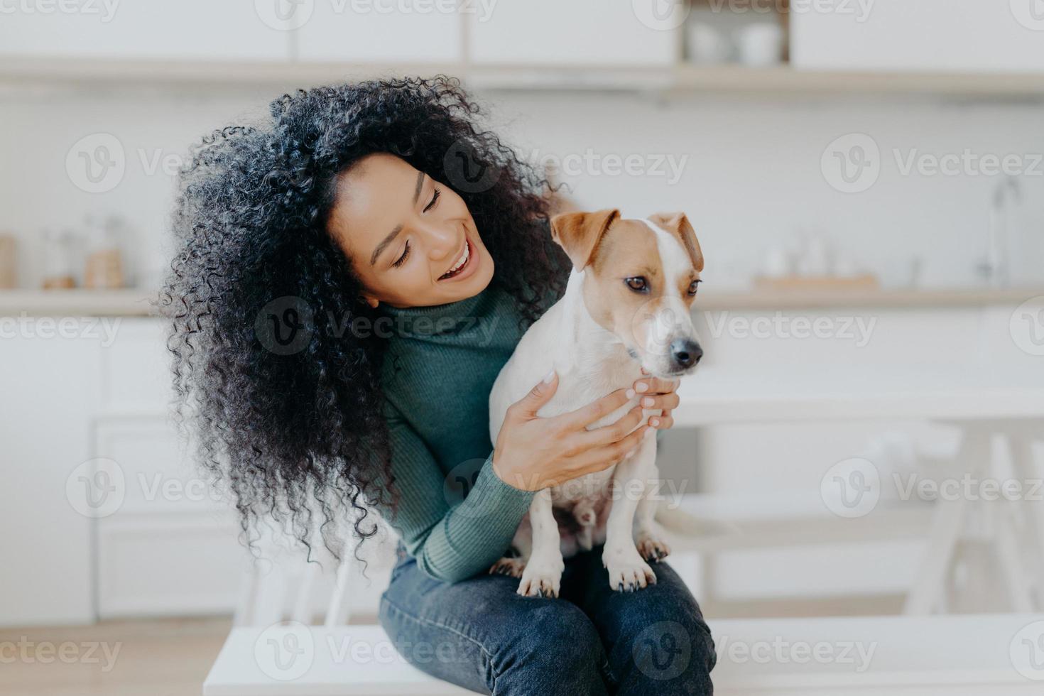 gelukkige vrouwelijke eigenaar van jack russell terrier-hond, voelt de verantwoordelijkheid om voor het huisdier te zorgen, heeft borstelig donker krullend haar, zit tegen een wazige keukenachtergrond. mensen en relatie met dieren foto