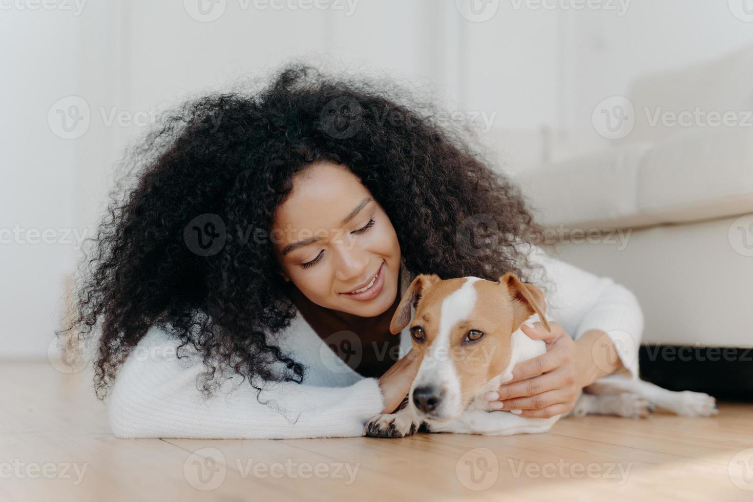 ontspannen afro-vrouw met helder donker haar ligt op de vloer, speelt met schattige puppy, heeft plezier met jack russell terrier hond draagt witte trui in de woonkamer. gelukkige eigenaar die lief huisdier aait foto