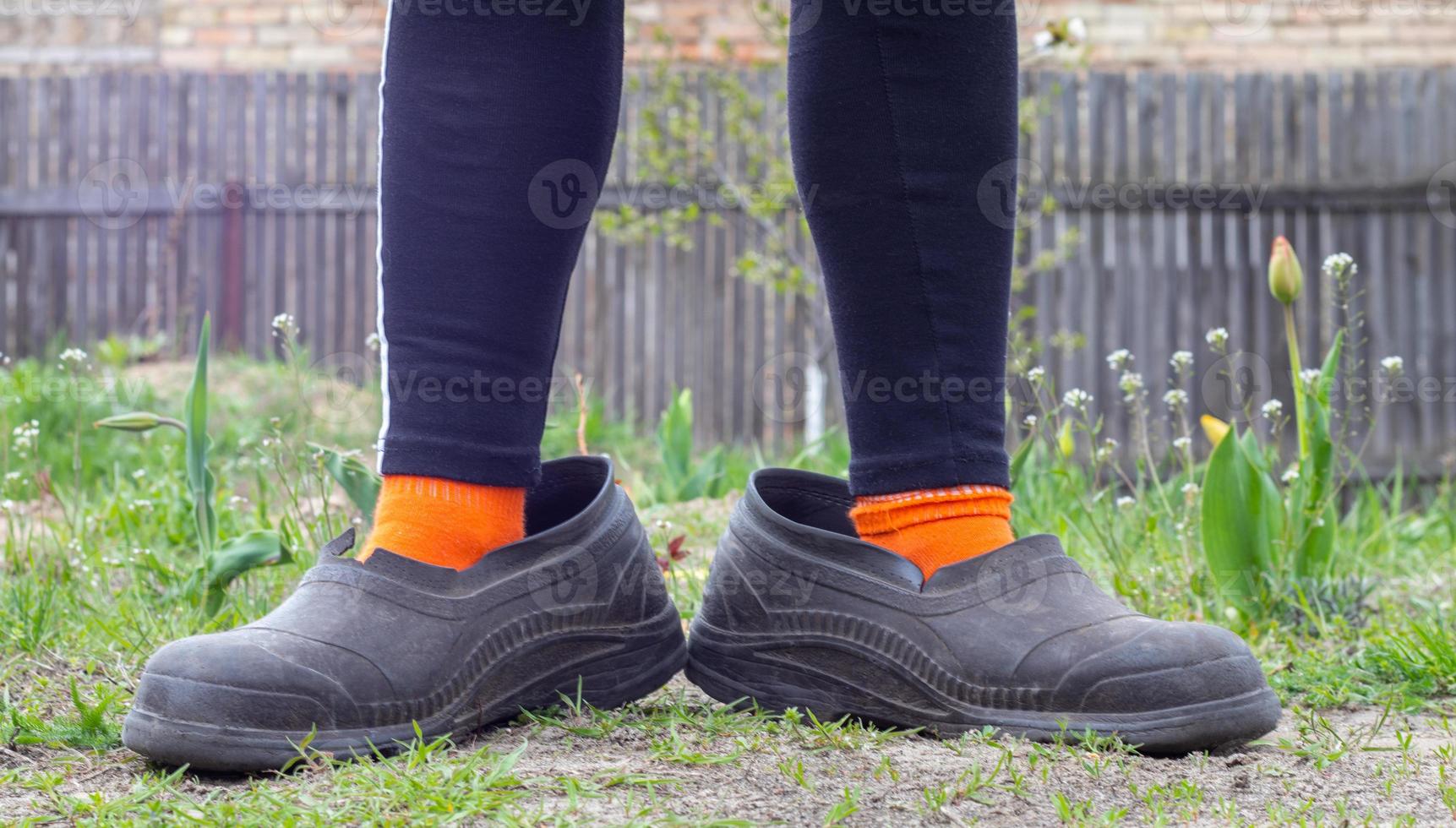 damesbenen in oversized rubberen overschoenen tegen de achtergrond van het platteland. benen in zwarte leggings, oranje sokken en vuile overschoenen in een buitenwijk. erg grappige foto. foto