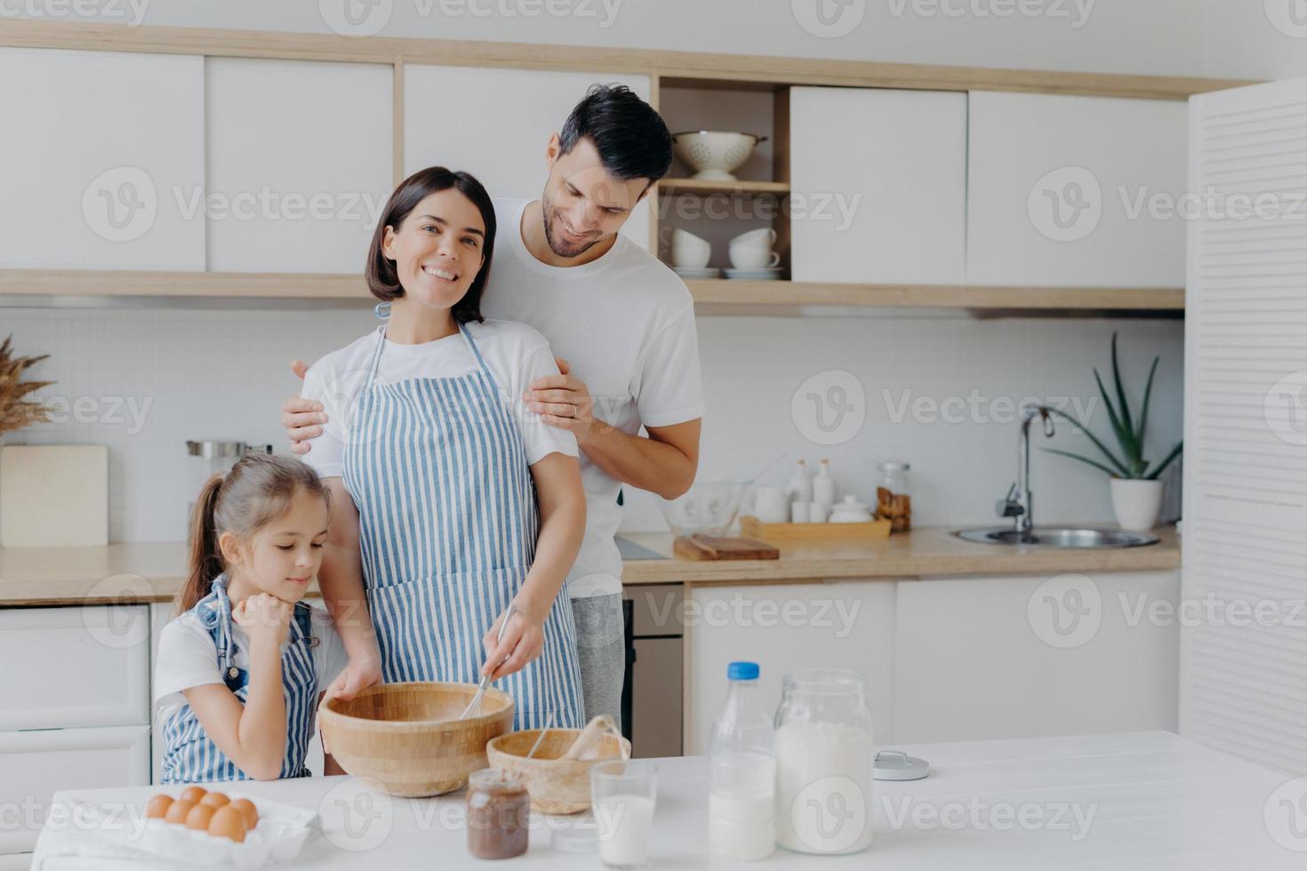 gelukkige familie koken samen in de keuken. vader, moeder en dochter druk bezig met het bereiden van een heerlijke maaltijd thuis. man omhelst vrouw die zwaait en deeg bereidt, koekjes bakt. eten, samenzijn foto