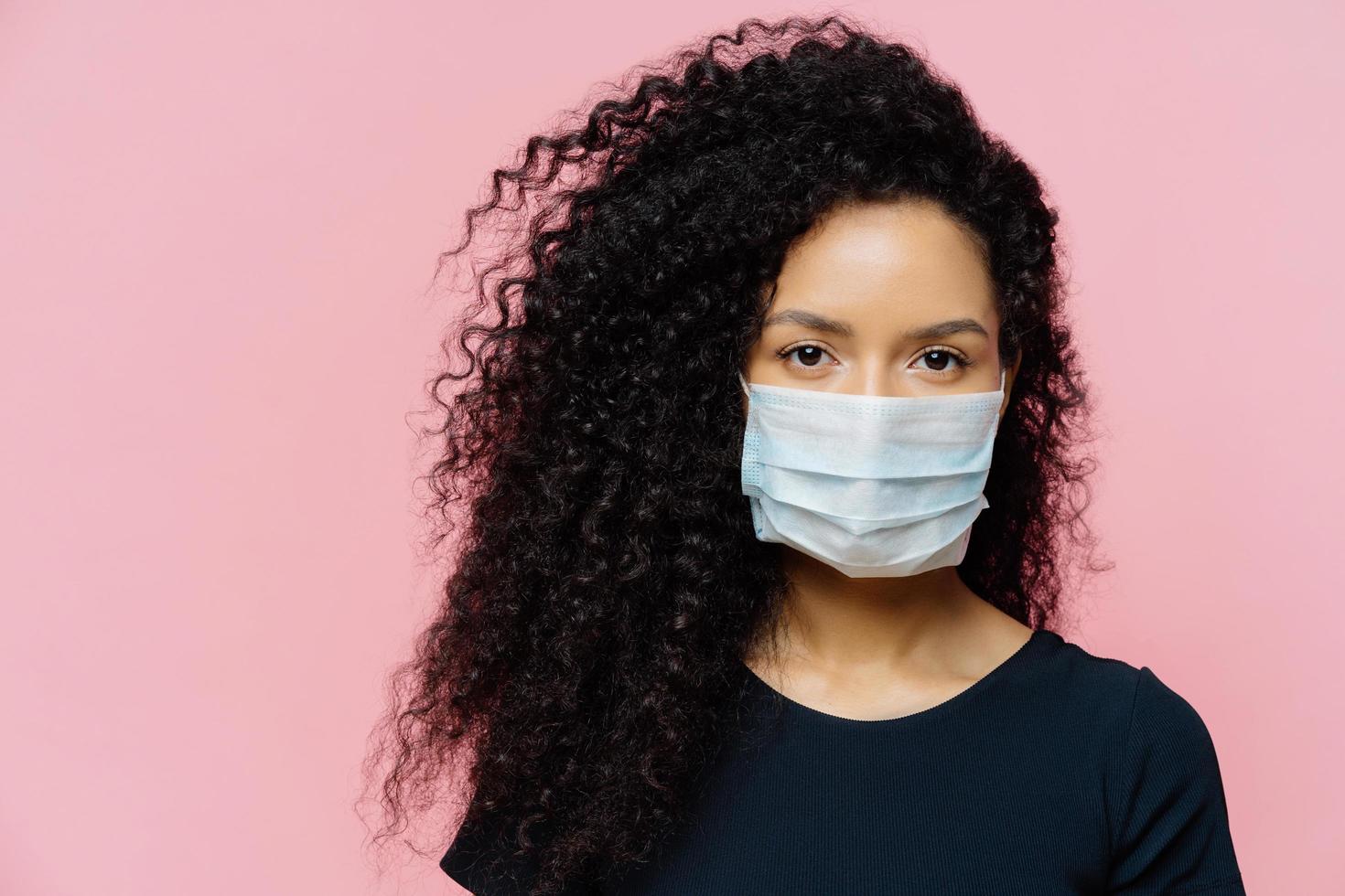 ernstige jonge vrouw met een donkere huid die thuis in zelfisolatie zit, een beschermend medisch masker draagt, thuis in quarantaine is, nonchalant gekleed is, poseert tegen een roze achtergrond. coronavirusziekte foto