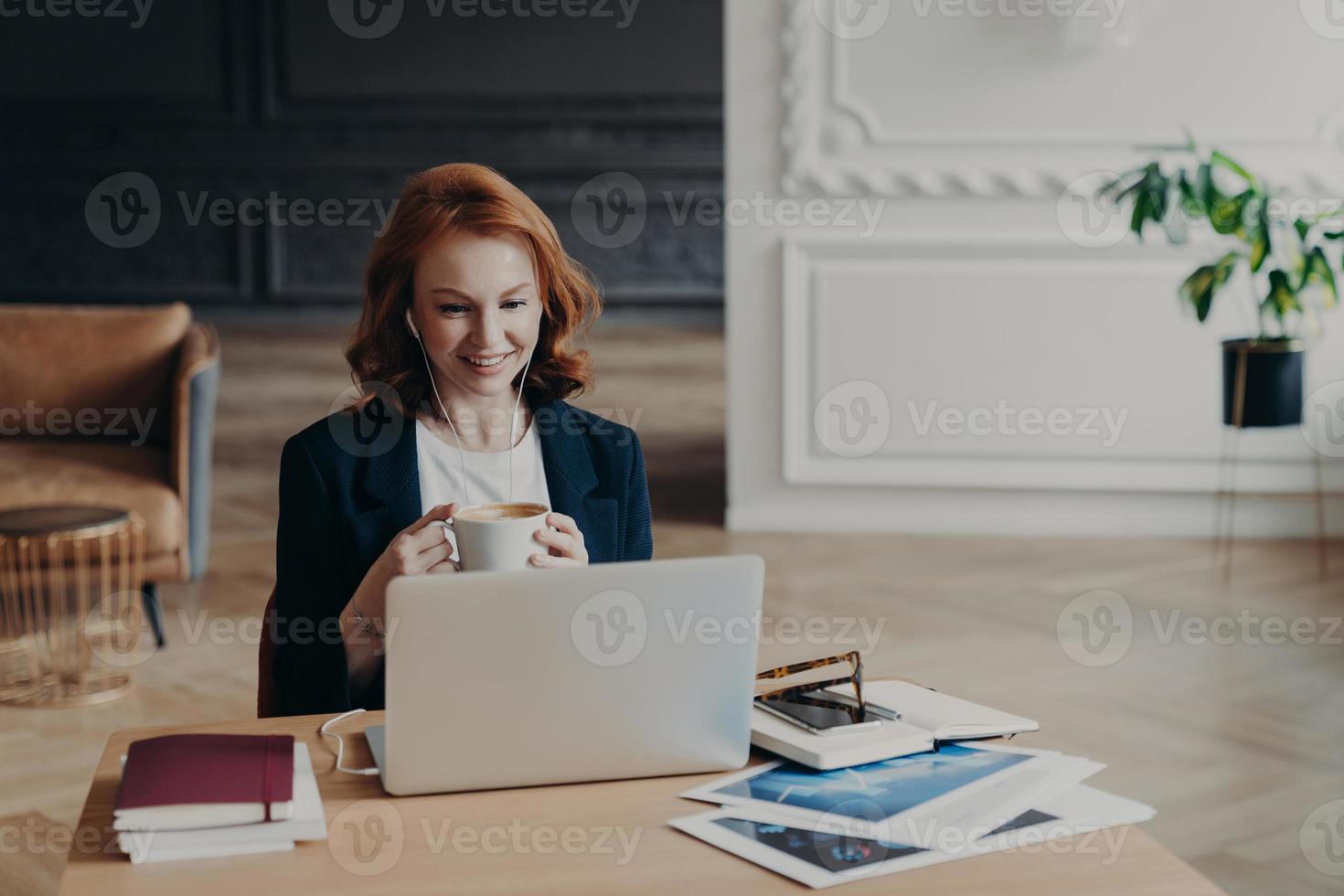 blije roodharige vrouw heeft een baan op afstand, communiceert in videochat met collega's, bespreekt ideeën voor project, geniet van online conferenties gebruikt moderne laptop en accessoires tevreden met vriendelijk praten foto