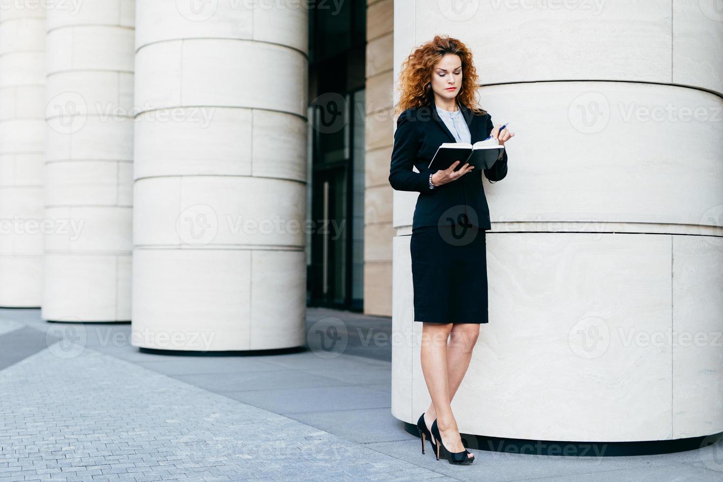 slanke jonge zakenvrouw in elegante kleding en schoenen met hoge hakken, met een geconcentreerde blik in haar notitieboekje, het vinden van een vrije datum voor een ontmoeting. serieuze vrouwelijke freelancer met haar dagboekboek en pen foto