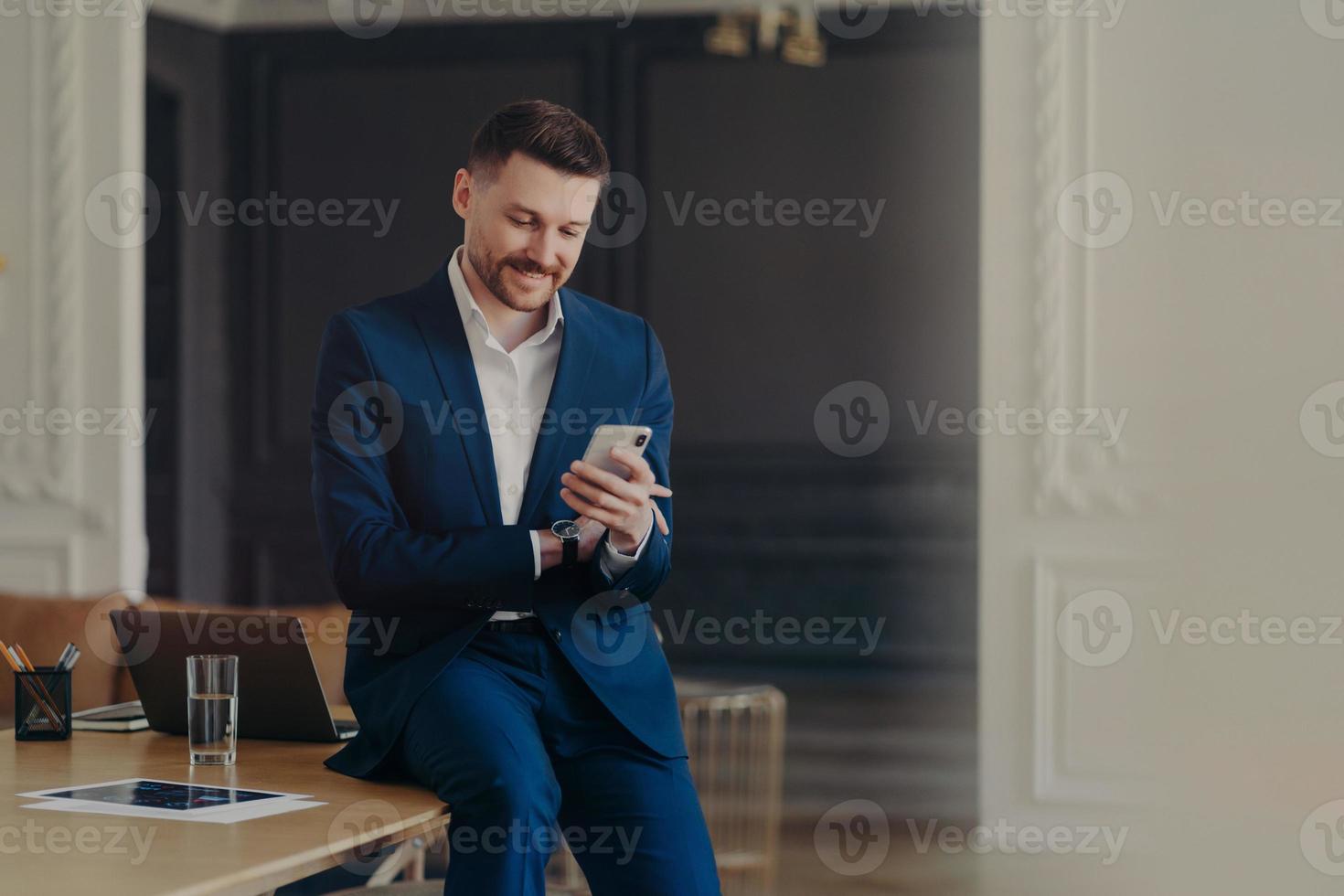 gelukkige knappe zakenman die naar mobiele telefoon kijkt terwijl hij op een bureau zit foto