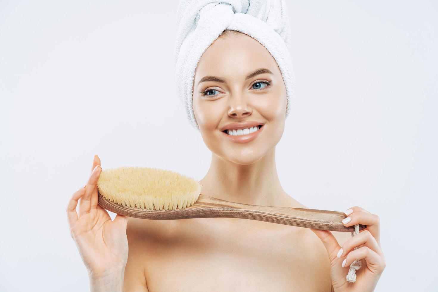 jonge vrouw met een gezonde, frisse huid, gebruikt een lichaamsborstel, glimlacht zachtjes, draagt een badhanddoek op het hoofd, poseert topless, geïsoleerd op een witte achtergrond. spa-accessoires. vrouwen, lichaamsverzorging, hygiëneconcept foto