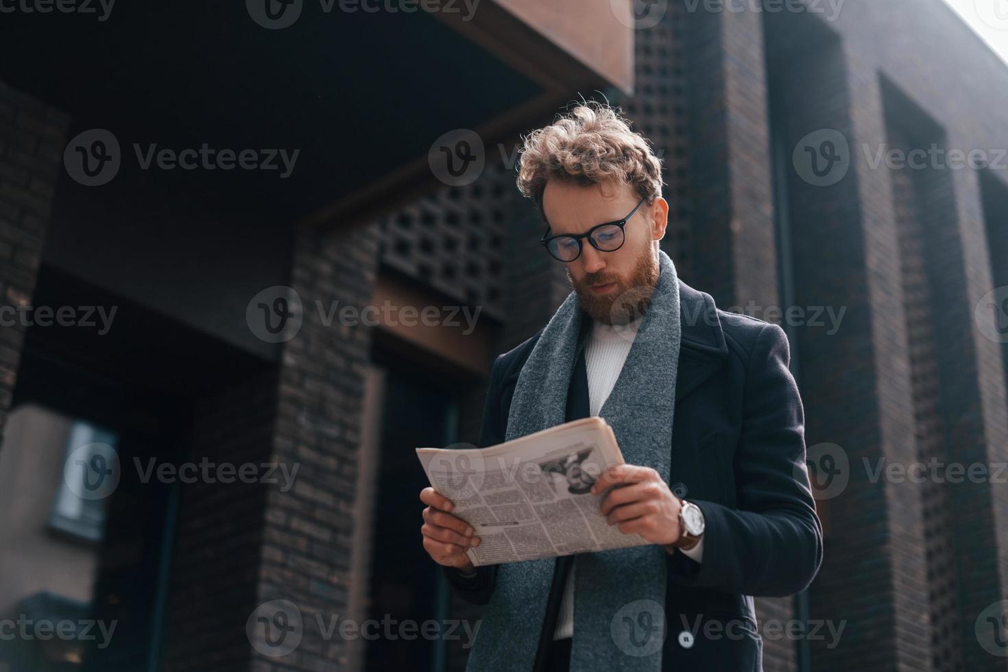 met krant in handen. stijlvolle man met baard en bril is buiten in de buurt van gebouw foto