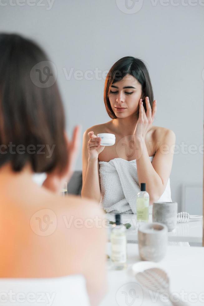 bij de spiegel zitten en crème op het gezicht gebruiken. jonge vrouw is thuis. conceptie van schoonheid foto