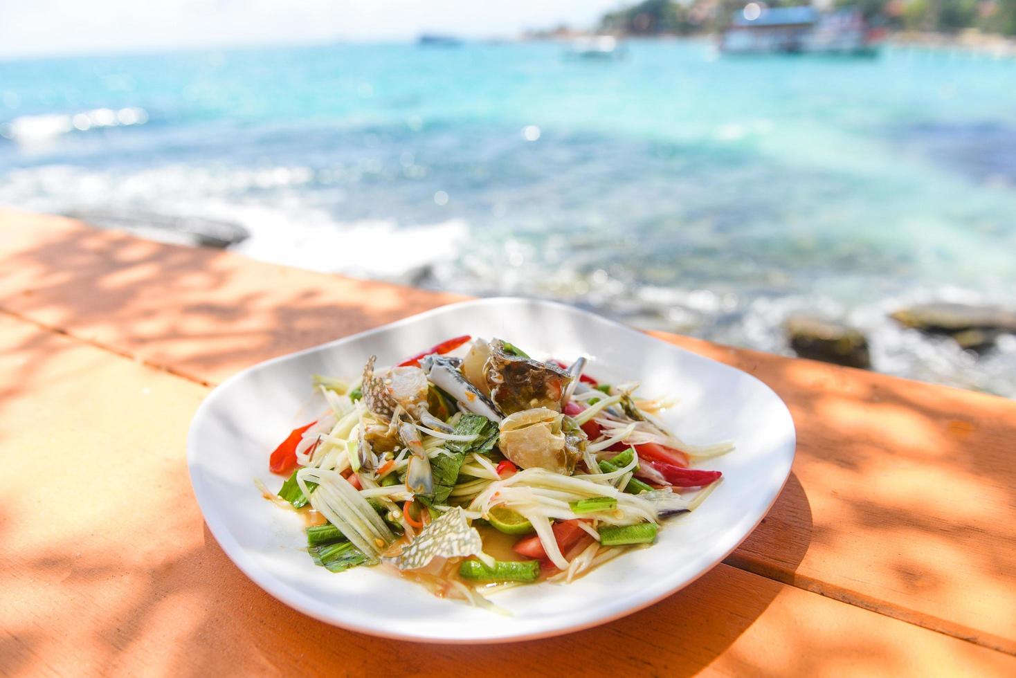 papajasalade met blauwe krab op tafel en strand zeekust achtergrond thais eten rauwe krab pittige salade zeevruchten en plantaardig voedsel op het zeeconcept foto