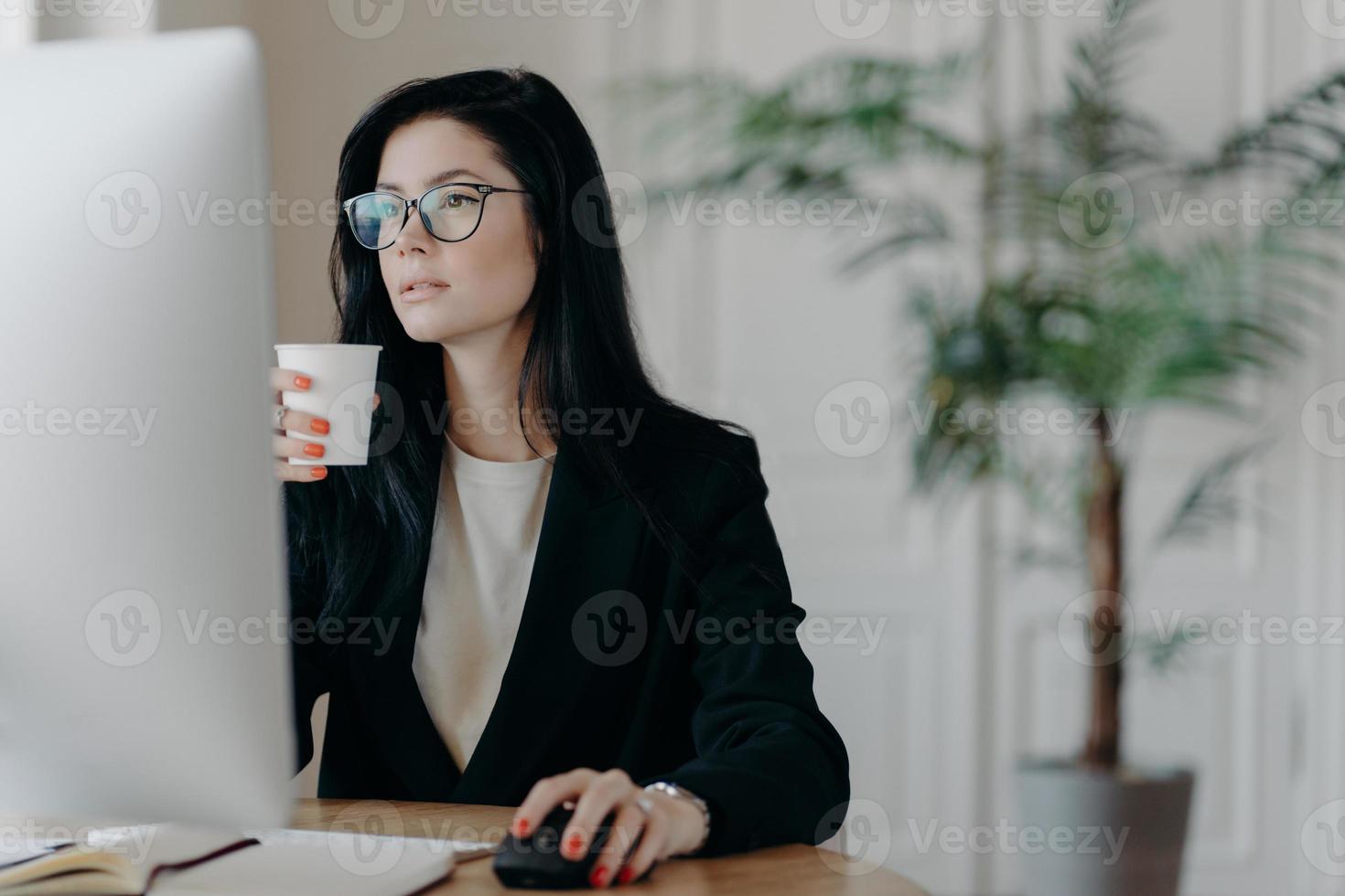 drukke jonge vrouwelijke administratieve poses op desktop, drinkt warme koffie uit papieren beker, formeel gekleed, ontwikkelt bedrijfsstrategie, plant opstarten, betrokken bij werkproces. bezettingsconcept foto