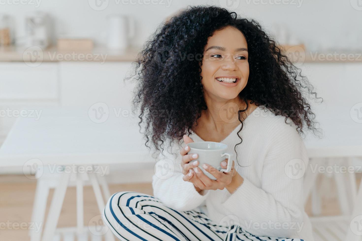 foto van schattige vrolijke vrouw met borstelig krullend haar kijkt weg met een glimlach, houdt mok koffie vast, draagt casual kleding, geniet van een gezellige rustige ochtend, proeft smaak. afro lag in de keuken tijdens vrije dag