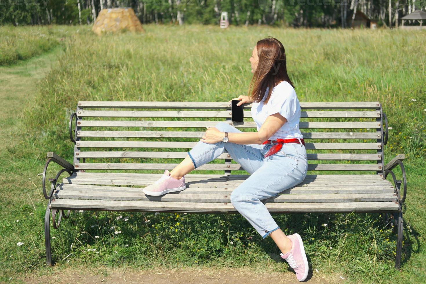 jonge mooie vrouw zittend op een bankje en genieten van aangenaam weer. zijaanzicht. vrouw die opzij kijkt en een smartphone vasthoudt. alleen reizen en lokaal reisconcept foto