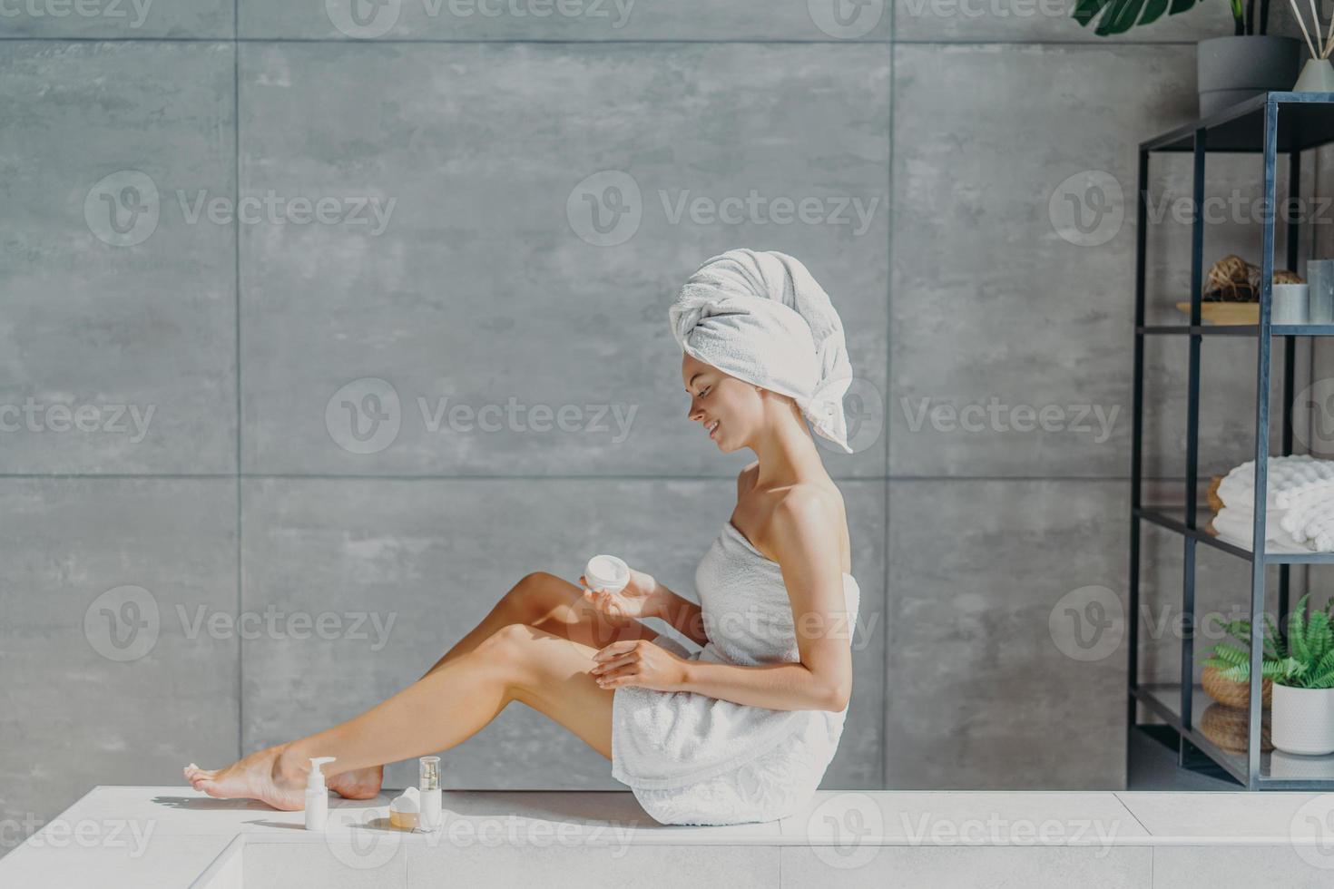 horizontaal schot van ontspannen jonge europese vrouw brengt vochtinbrengende lichaamscrème aan op benen, gewikkeld in badhanddoek, heeft een tedere glimlach, gezonde verfrist huid na het nemen van een bad, poseert in een gezellige badkamer. foto