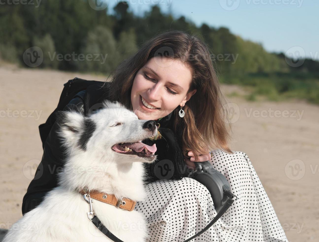gelukkige laika-hond lacht en zit naast zijn eigenaar. jonge mooie vrouw streelde haar hond zittend op een zandstrand. saamhorigheid en liefdevol huisdierenconcept, huisdierenliefde foto