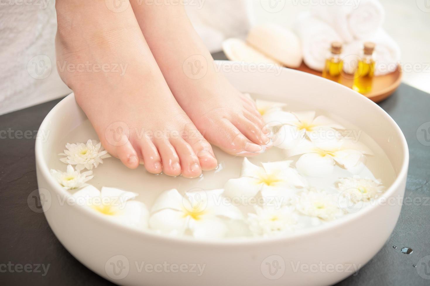 close-upmening van vrouw die haar voeten in schotel met water en bloemen op houten vloer weken. spa-behandeling en product voor vrouwelijke voeten en hand spa. witte bloemen in keramische kom. foto