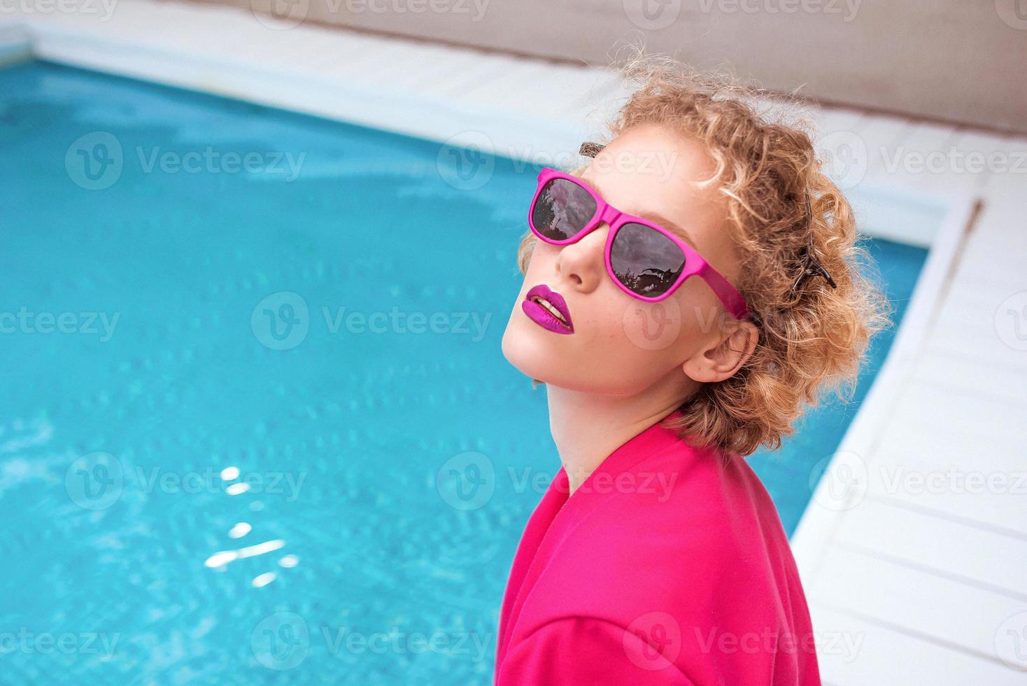 portret van prachtige stijlvolle roodharige krullende jonge vrouw zittend in fuchsia jas, stijlvolle zonnebril en zwarte onderbroek bij het zwembad. foto