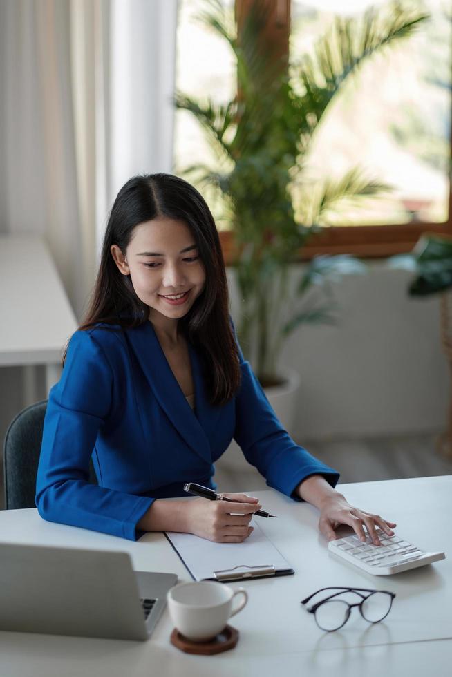 portret jonge Aziatische zakenvrouw zittend aan hun bureau en notities maken met behulp van laptop en rekenmachine op kantoor. foto