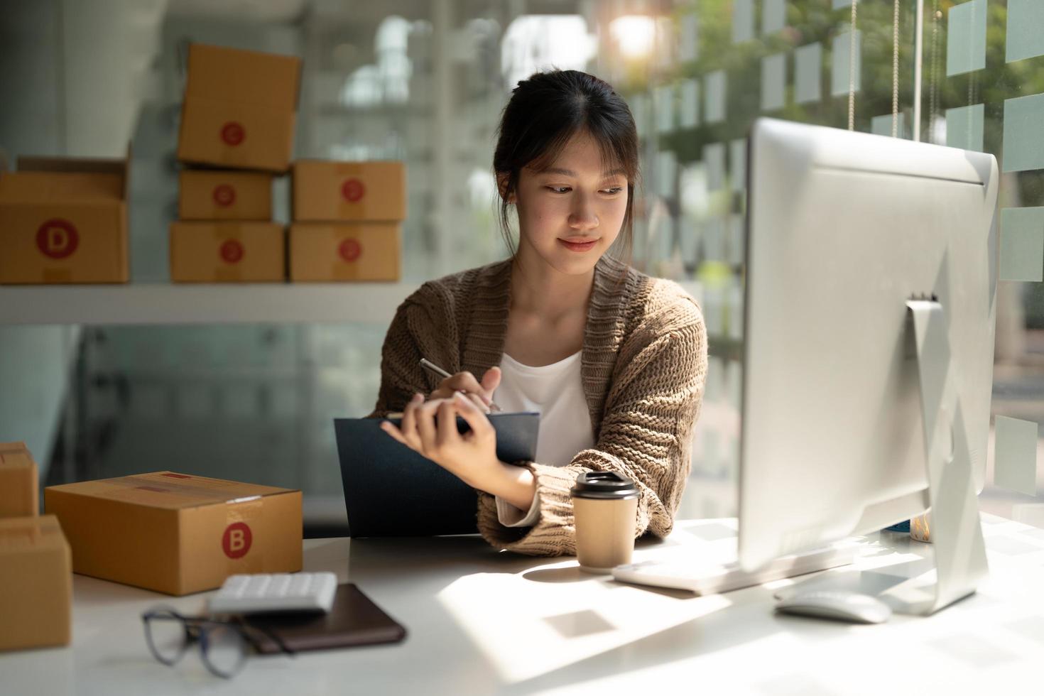 jonge vrouw kleine ondernemer online winkelen thuis. lijst bestellingen van klanten met mobiele telefoons die pakketproducten op de achtergrond voorbereiden. MKB ondernemer of freelance levensstijlconcept. foto