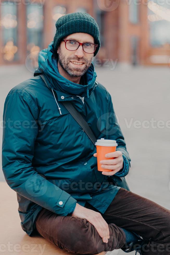 stijlvolle blanke man heeft vrije tijd, poseert buiten met wegwerpkop koffie, gekleed in hoed en jas, geniet van goede rust, wacht op iemand op straat, heeft een tevreden uitdrukking. levensstijl concept foto