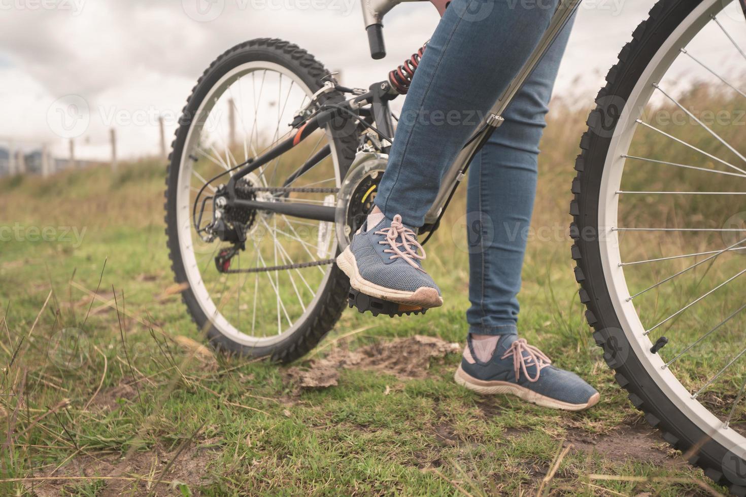 detail van de voeten van een jonge vrouw die een blauwe broek draagt op de pedalen van haar fiets in het midden van het veld op een bewolkte dag foto