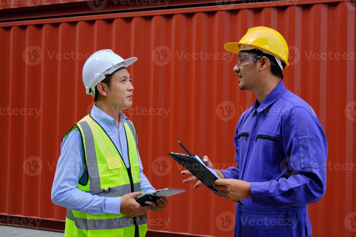 twee professionele aziatische mannelijke arbeiders in veiligheidsuniformen en veiligheidshelmen op een stalen containerachtergrond, werken op de logistieke terminal, laden controleverzendingsgoederen voor de vrachtvervoersindustrie. foto