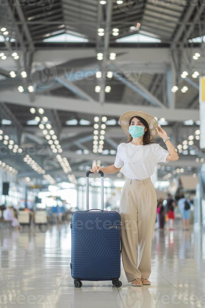 jonge vrouw met gezichtsmasker met bagage die op de luchthaven loopt, bescherming tegen infectie met coronavirusziekte, aziatische vrouwelijke reiziger met hoed. tijd om te reizen na het concept van de boosterdosis van het vaccin foto