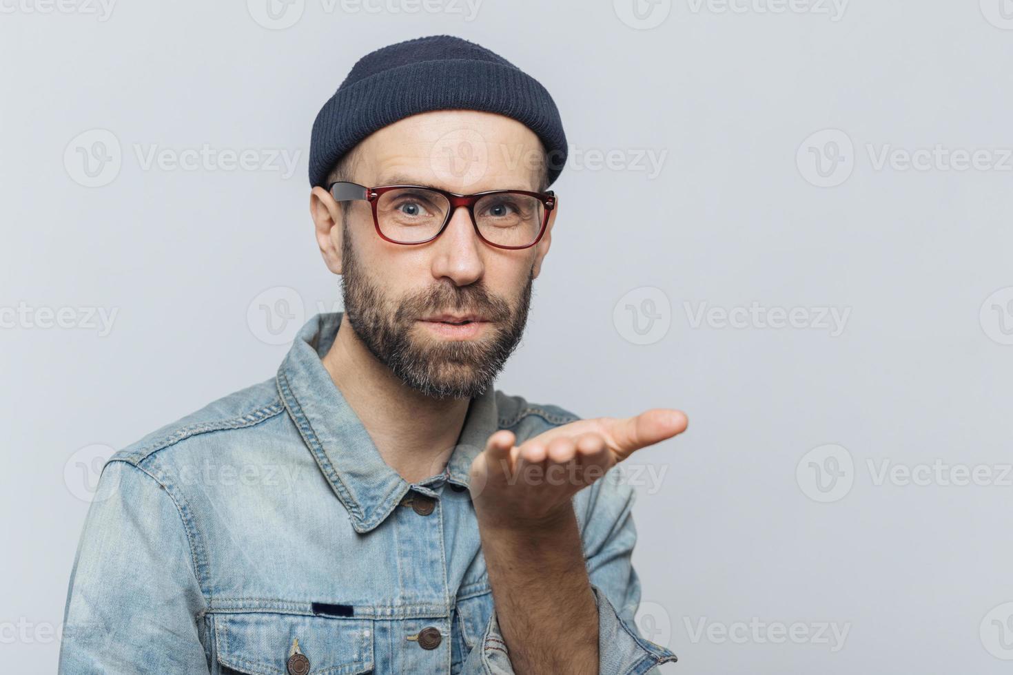 aangenaam uitziende jonge bebaarde man met snor, draagt modieuze bril, zwarte hoed en denim shirt, blaast luchtkus op camera, drukt zijn liefde uit aan vriendin, geïsoleerd over grijze achtergrond foto