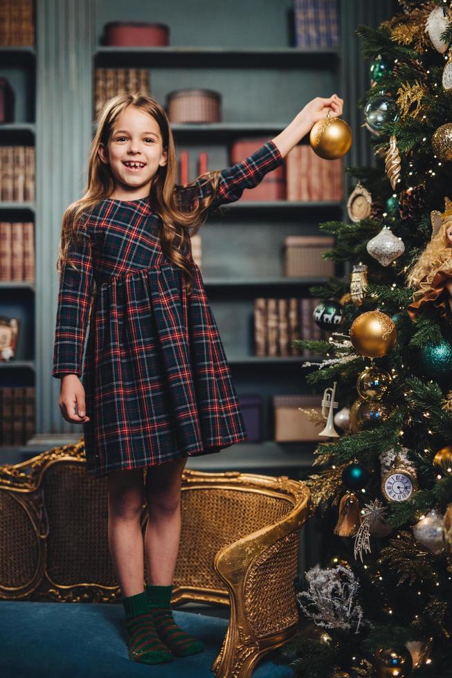 glimlachend mooi klein meisje staat op een fauteuil, houdt een glazen bol vast, versiert de nieuwjaarsboom, is blij om het te doen. schattig klein kind bezig met versiering van de kerstboom. seizoen en feest foto