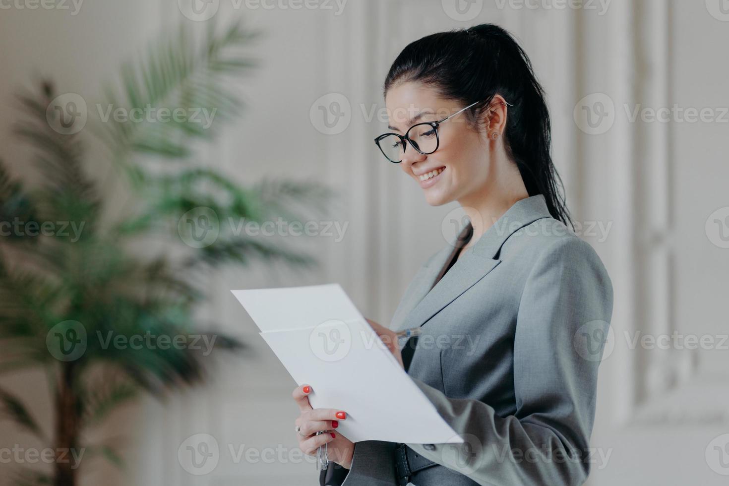 blije vrouwelijke advocaat schrijft in zakelijke papieren documenten, draagt een optische bril, formele kleding, staat in eigen kast, ontwikkelt nieuwe strategie, heeft een gelukkige uitdrukking. mensen, carrière, werkgelegenheidsconcept foto