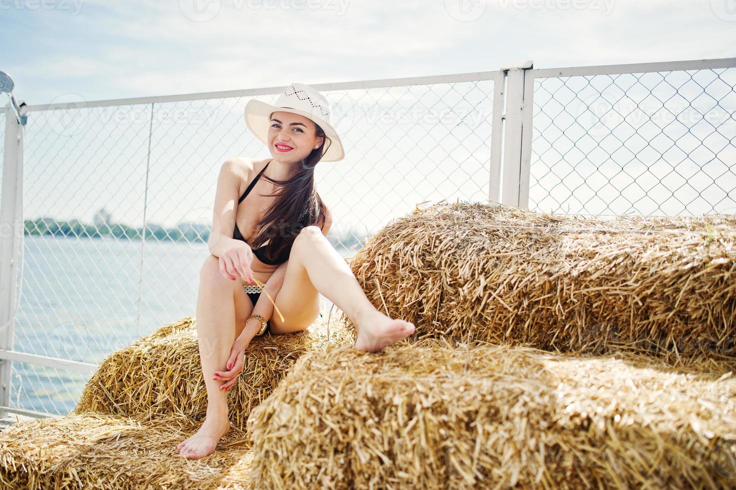 portret van een prachtig meisje in zwarte bikini zwembroek poseren op de hooibaal met een hoed aan het meer. foto