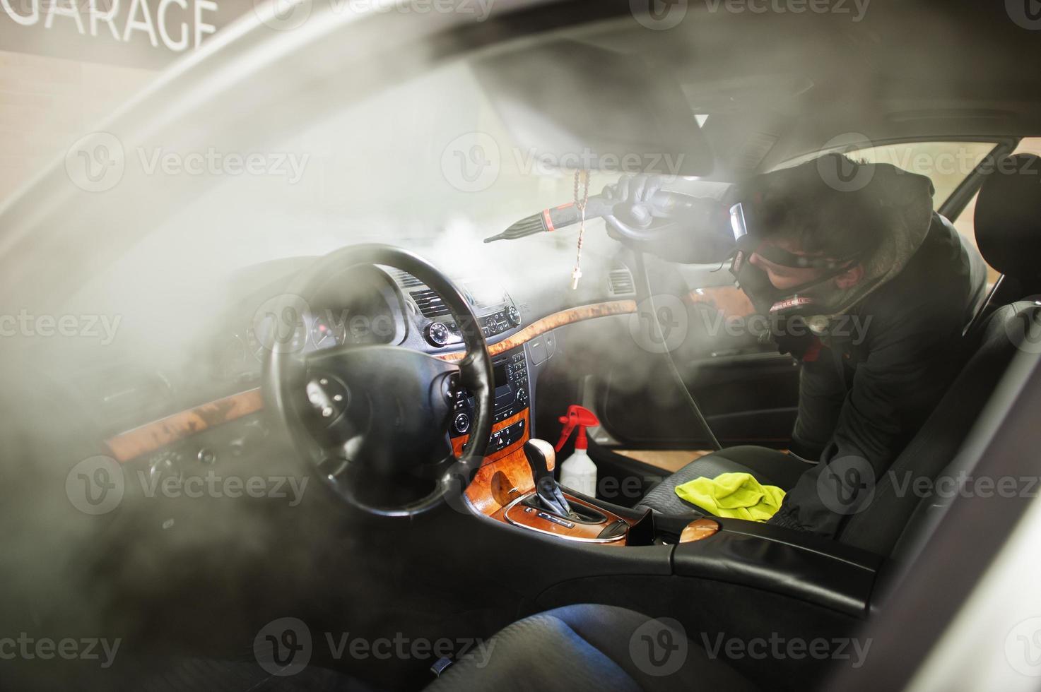 man in uniform en gasmasker, werknemer van autowascentrum, auto-interieur schoonmaken met hete stoomreiniger. auto detaillering concept. foto