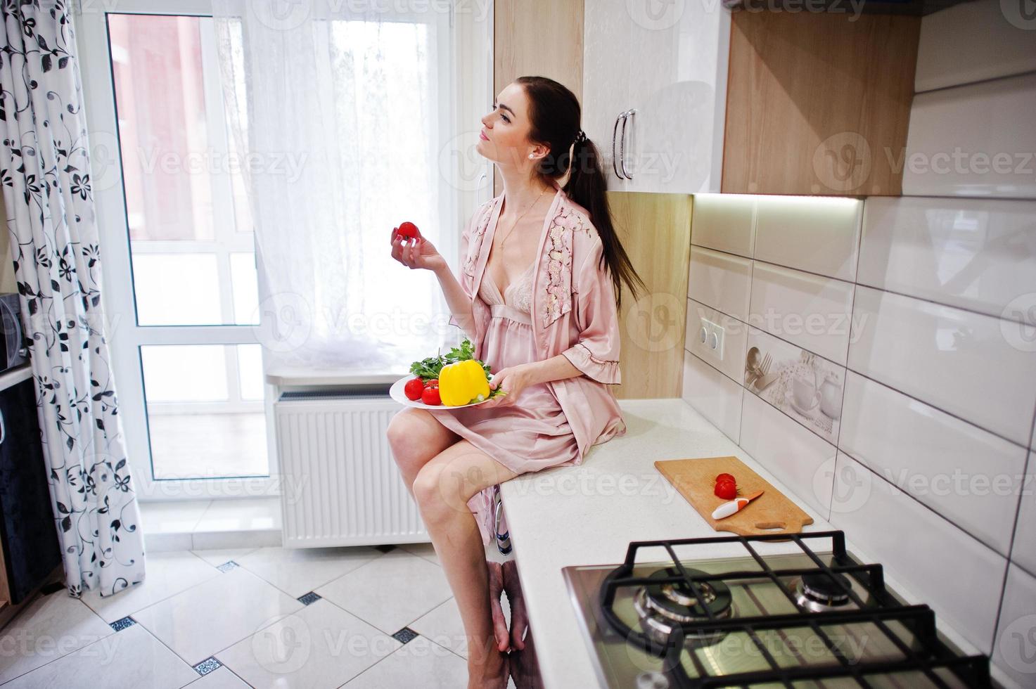 portret van een mooie jonge vrouw in roze gewaad poseren met een tomaat in haar keuken. foto